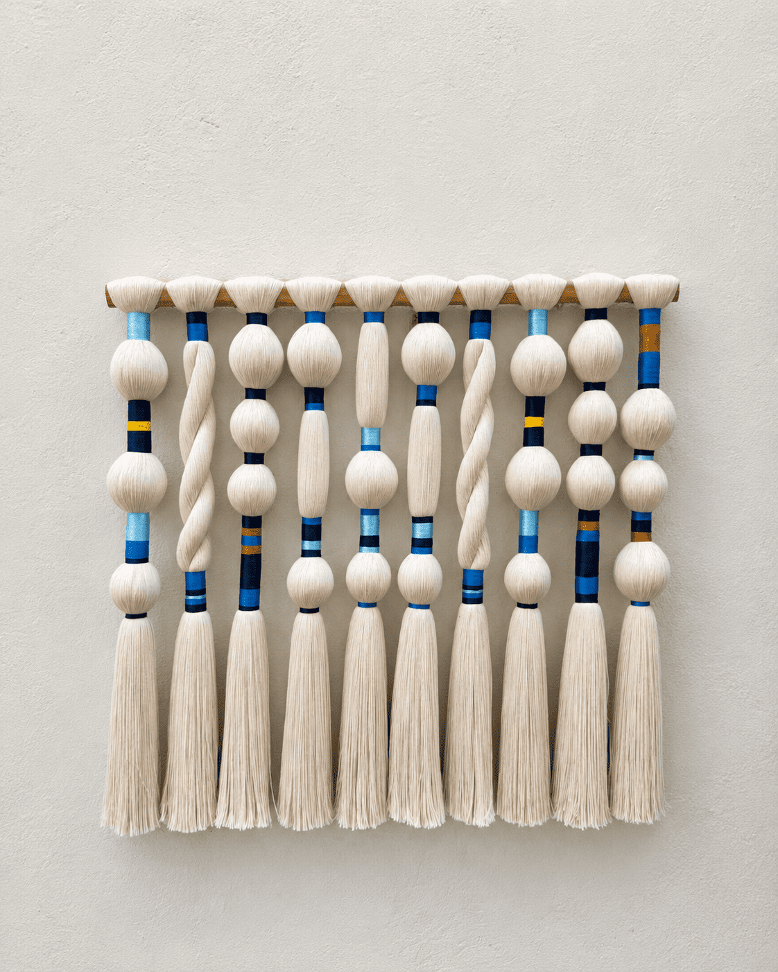 Painel Gaia escultural com onze colunas de fios creme e detalhes em azul e amarelo sobre suporte de madeira