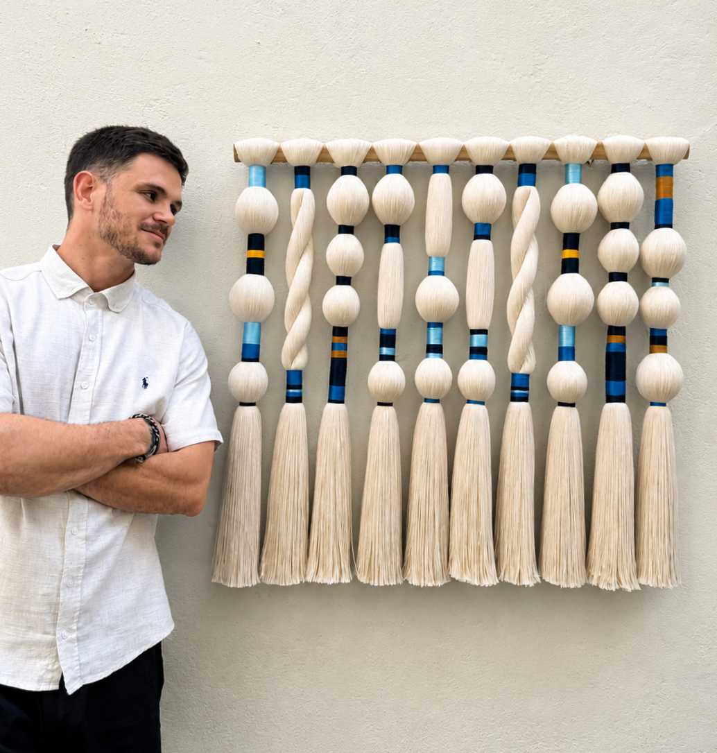 Giovanni Zorzal ao lado de painel escultural de macramê com fios creme e detalhes coloridos em azul e amarelo